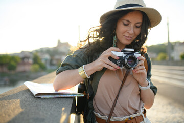 Young beautiful female traveler with map standing on bridge and using camera on sunny day in city.