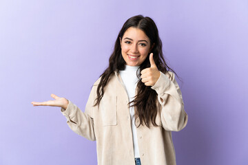Wall Mural - Young caucasian woman isolated on purple background holding copyspace imaginary on the palm to insert an ad and with thumbs up