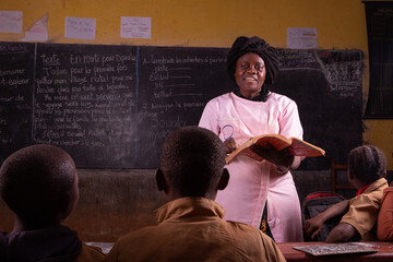 black female teacher stands in front, explains the lesson by reading from the book from the desk