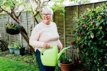 Portrait Of Happy, Smiling Senior, Mature Woman Watering Flowers With Watering Can In The Garden In A Sunny Day. Retired Elderly Woman And Gardening Hobby. Active Retirement Lifestyle.