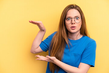 Young caucasian woman isolated on yellow background shocked and amazed holding a copy space between hands.