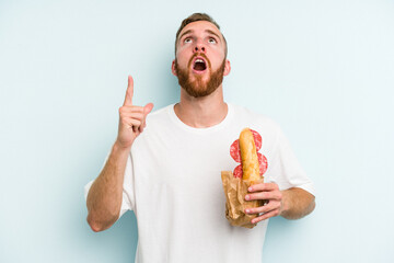 Young caucasian man eating a sandwich isolated on blue background pointing upside with opened mouth.