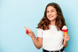Little caucasian girl eating an ice cream isolated on blue background smiling and pointing aside, showing something at blank space.