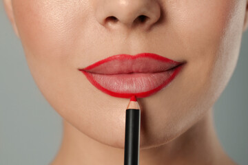 Young woman applying beautiful red lip pencil on grey background, closeup