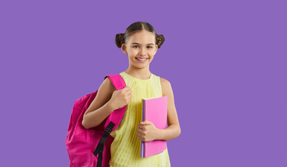 Studio shot of happy school child with books and backpack. Cheerful beautiful girl in yellow top standing isolated on purple background, holding pink bag and notebooks, looking at camera and smiling