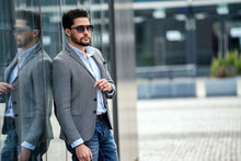 Outdoor Photo Of Handsome Elegant Man Wearing Fashionable Clothes And Sunglasses, Posing On The Wall With Reflections. City Stale. Male Model.