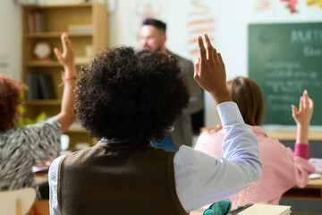 Rear view of African American adolescent guy raising hand to answer question of teacher while sitting in front of his classmates