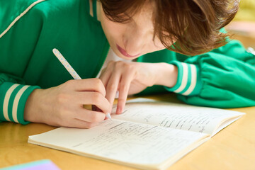 Wall Mural - Teenage girl in green jacket bending over desk while making notes in copybook or writing down lecture after teacher at lesson