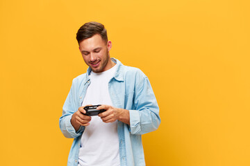 Overjoyed tanned handsome gamer man in blue basic t-shirt hold joystick look at gamepad posing isolated on orange yellow studio background. Copy space Banner Mockup. People lifestyle concept