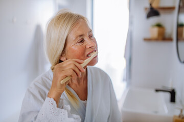 Poster - Beautiful senior woman in bathrobe brushing teeth with eco wooden toothbrush inbathroom, sustainable lifestyle.