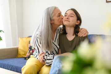 Wall Mural - Senior grandmother with teenage granddaguhter having good time together at home, hugging and kissing on cheeck.