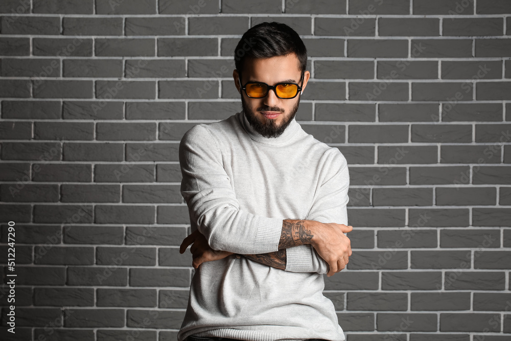 Young bearded man with crossed hands wearing sunglasses on grey brick wall background