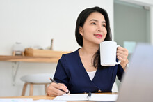 Successful Young Asian Businesswoman At Her Office Desk Sipping Morning Coffee.