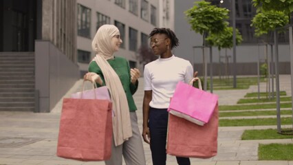 Wall Mural - Two happy satisfied multiethnical girls, walking down the old city street after shopping, carrying colorful bags in their hands, smiling at camera. Shopping in the city, urban lifestyle.