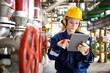 Refinery worker with hardhat and earmuffs checking parameters on tablet computer for oil and gas production.