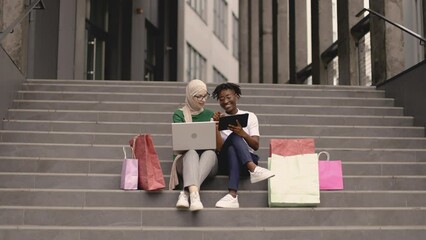 Wall Mural - Outdoor full length fashion portrait of two young multiracial ladies in stylish clothes sitting on the steps of a large shopping mall and ordering clothes online colored shopping bags lie next to them