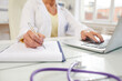 © Mediaphotos - Close-up of unrecognizable doctor sitting at table and using laptop while filling document with medical history of patient