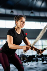 Powerful woman training battle ropes at cardio workout in dark gym. Professional athlete exercise fitness sport club equipment. Strong bodybuilder lifting weights. Athletic person effort