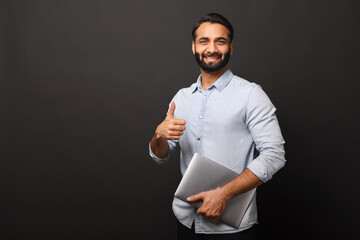 Indian male entrepreneur in casual shirt carrying laptop and showing thumb up isolated on black, bearded businessman with laptop looking at the camera and smiles, ok gesture