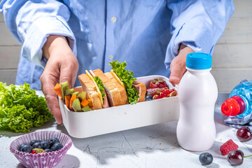 Back to school. Mother female hands making kids school lunch box with fresh fruits, berry, yogurt, healthy sandwich, vegetable snacks. White kitchen marble table copy