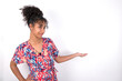 © Jihan - Portrait of Young African American woman wearing colourful dress over white wall  with arm out in a welcoming gesture.