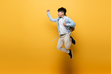 Wall Mural - Side view of asian schoolkid with backpack jumping on yellow background.