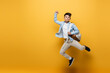 Asian schoolkid holding backpack and showing yes gesture while jumping on yellow background.