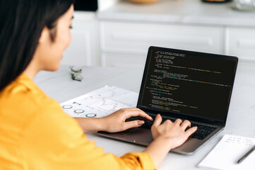 Over shoulder view of asian woman, IT specialist, at a laptop with programming code on the screen. Smart professional female programmer working with a software and coding technology, writing codes