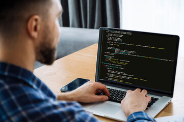 Developing coding technologies, IT concept. Over shoulder view of male IT-specialist to a laptop with programming code on the screen. Smart male programmer working on new software application
