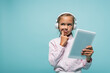 Pensive schoolkid in headphones holding digital tablet isolated on blue.