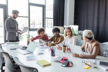 man in eyeglasses standing with laptop near multicultural team talking in advertising agency.