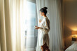 © Alla - Young woman wearing white bathrobe opening curtains in luxury hotel room and enjoying sun in the morning while having her cup of coffee