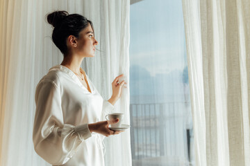 attractive woman is holding a cup with hot tea or coffee and looking at the sunrise while standing n