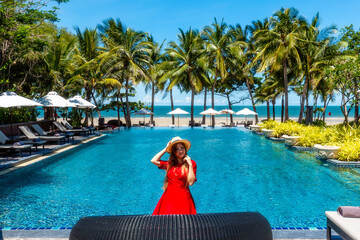 Travel vacation woman in red dress enjoying a summer vacation near swimming pool in tropical resort near the beach with sea view. Carefree girl tourist in hat relax in luxury resort, Thailand