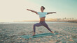 Fit woman doing yoga on mat by the sea. Beautiful yogi girl in sportswear standing in warrior yoga pose on the beach