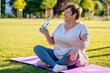 Relaxed senior plus size woman with earphones sitting on yoga mat on green grass outdoors resting after exercises drinking water at warm sunny summer day