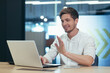 © Liubomir - Working video call in the office. Young male businessman, freelancer sitting at a desk in the office on a laptop, talking through the camera on a video call, explaining something, waving his hands.