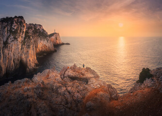 Wall Mural - Aerial view of man on mountain peak, lighthouse, sea at colorful sunset in summer. Landscape with guy, high rocks, sea bay, stones, beach, orange sky. Top view of cliffs. Cape Lefkada, Greece. Nature