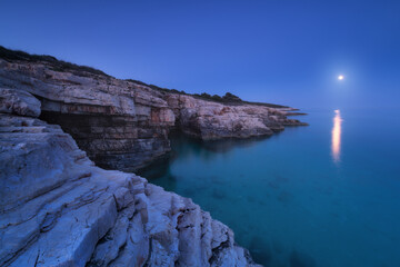 Wall Mural - Rocky sea coast and full moon at night in summer. Beautiful landscape with beach, cliffs, stones in blurred water, cave, blue sky at twilight. Nature. Adriatic sea at night, Kamenjak, Croatia. Travel