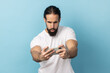 © khosrork - Portrait of bearded man wearing white T-shirt standing, using smartphone and playing mobile game with concentrated excited face. Indoor studio shot isolated on blue background.