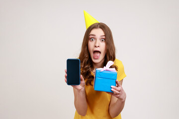 Wall Mural - Unbelievable birthday bonus for mobile user. Portrait of shocked teenage girl in birthday cone holding gift box and mobile cellphone with blank display. Indoor studio shot isolated on gray background.