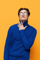 Wall Mural - a handsome man stands in black glasses for vision in a blue zip-up jacket, posing on an orange background with his hand on his neck