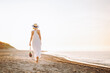 © Olga Rolencino - Back view of slim woman wearing long white light summer sundress, straw hat with black ribbon, walking on sandy beach near water sea on sunny day, carrying string bag full of products vegetables.
