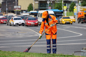 Wall Mural - Woman worker in orange uniform with a broom sweeps the road on cars background. Traffic jam, street cleaning in summer city