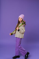 Portrait of a cute teenage girl with long hair and in a striped sweatshirt and lilac hat on her head, child holding a bouquet purple flowers, standing full-length on a purple background in the studio