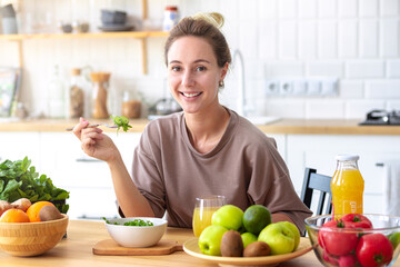 Wall Mural - Healthy lifestyle, diet, weight loss concept Beautiful happy woman eats breakfast looking at the camera and smiling friendly Beautiful female eating fresh salad while sitting in kitchen Vegan meal