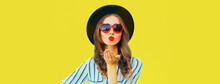 Portrait Of Beautiful Young Woman Blowing Her Lips Sending Air Kiss Wearing Red Heart Shaped Sunglasses, Black Round Hat, Striped T-shirt On Yellow Background, Blank Copy Space For Advertising Text