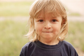 Wall Mural - close-up portrait of the pensive face of a little blond boy on a green background summer street