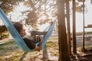 Wall Mural - Attractive young man resting in hammock with coffee, tea in hammock in forest. Rear.