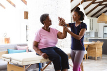 African American Female Physiotherapist Stretching Senior Woman's Wrist Sitting On Table At Home